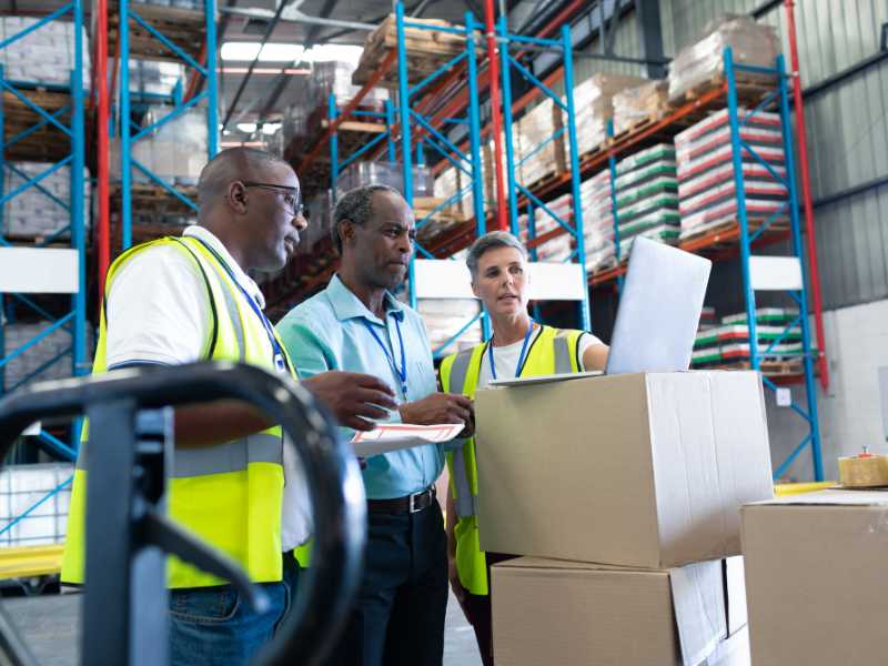 AMS Fulfillment workers looking at a lapton screen, which is sitting on a stack of boxes. 	