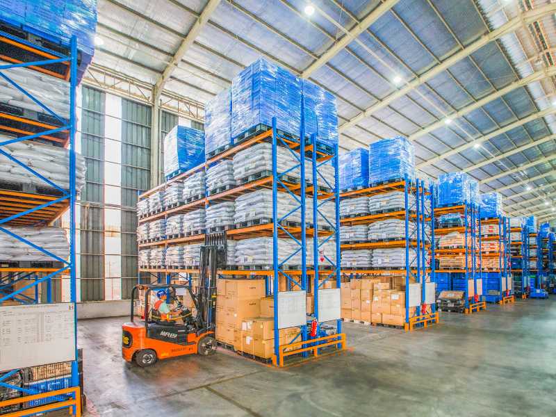 Shelves in a warehouse full of products, and a person in a forklift preparing to get a palette. 	