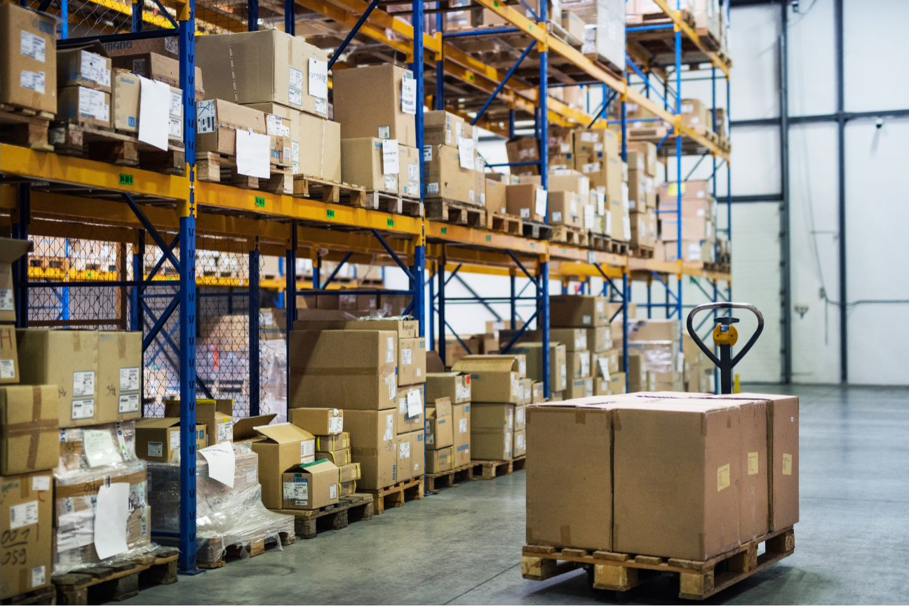 Image of a palet with packages, with shelves in the background stocked with packages. 