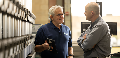A person wearing a blue shirt and holding a mini tablet talking to a person wearing a grey shirt.