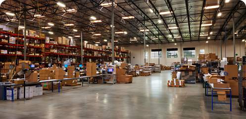 AMS warehouse showing boxes, assembly tables with blue legs, and red shelves filled with orders in the background.
