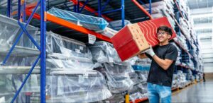A person is in a warehouse full of mattresses, holding a mattress in a box and smiling.