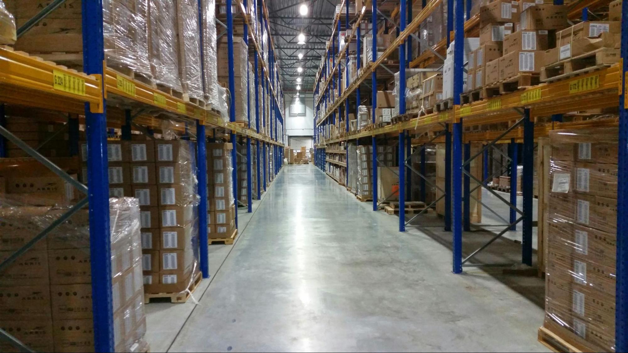 A long, well-lit warehouse aisle with tall blue and yellow metal shelving units fully stocked with palletized cardboard boxes and products wrapped in plastic.
