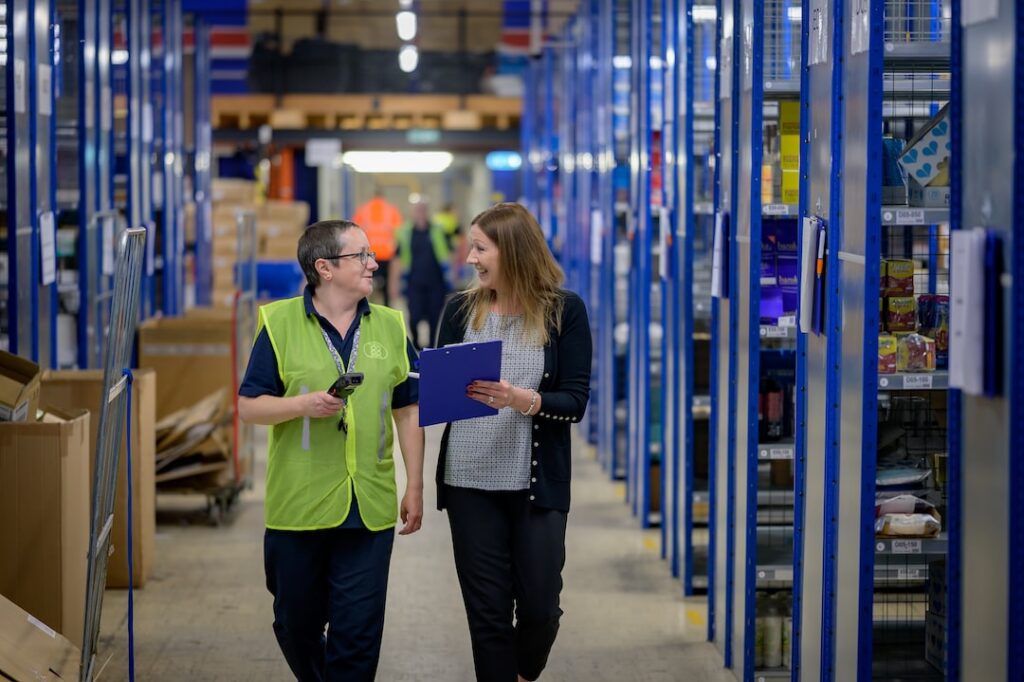 Two people are walking down a warehouse aisle filled with blue shelving, looking at each other and laughing.
