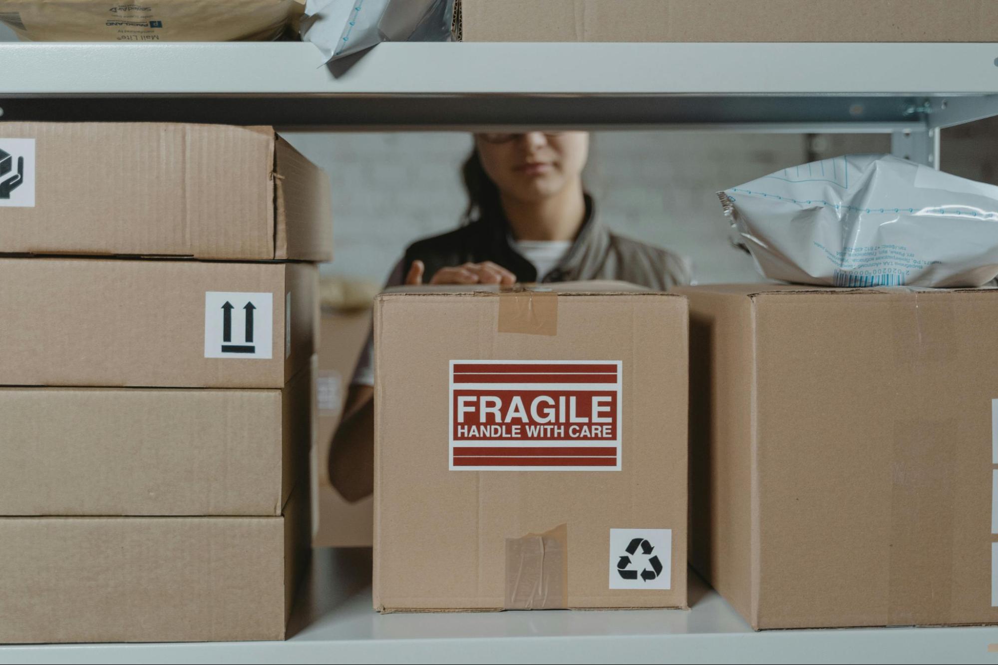 A person is placing a box on a shelf, with a red ‘Fragile, Handle With Care’ Sticker.