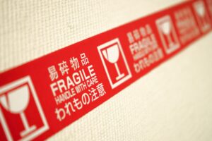 A red roll of shipping tape with white text and symbols, the text reads ‘FRAGILE’ and ‘HANDLE WITH CARE’ in English, as well as other languages.