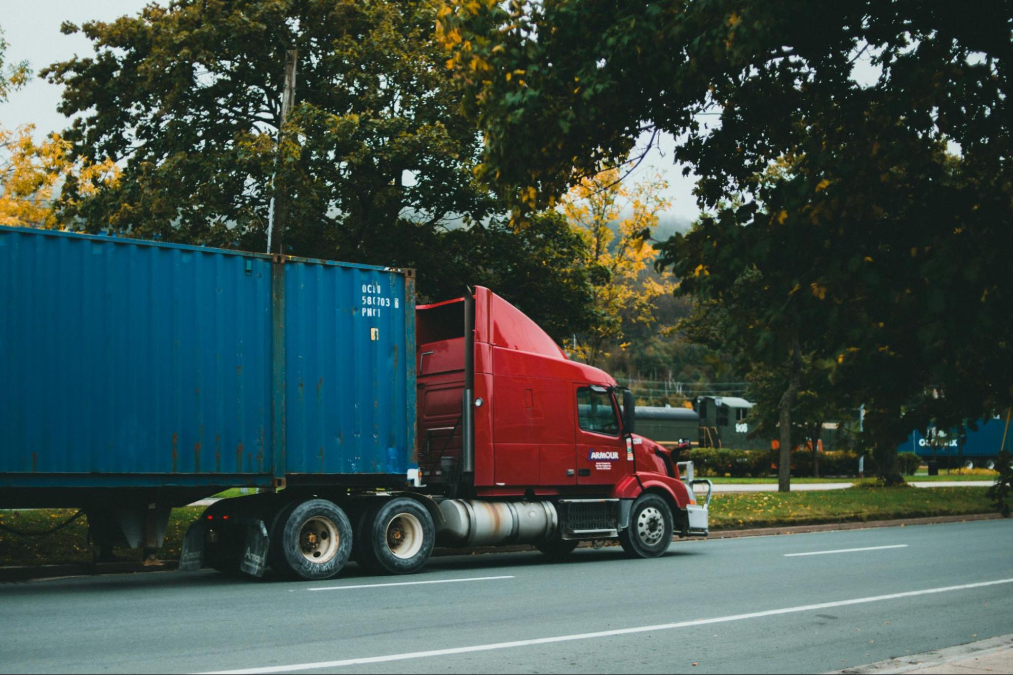 A red semi-truck is pulling a blue shipping container on a paved road, surrounded by trees.