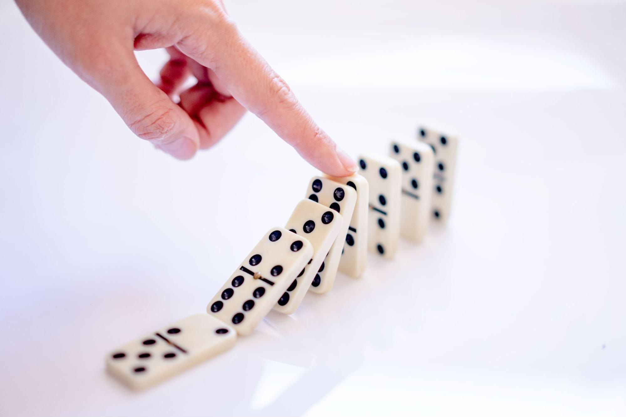 A finger pushing the first in a row of standing dominoes, triggering a chain reaction.