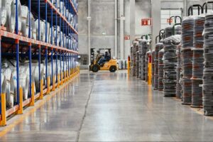 A person is driving a yellow forklift through a warehouse, with large coils of wire that are wrapped in plastic on either side of the aisle.
