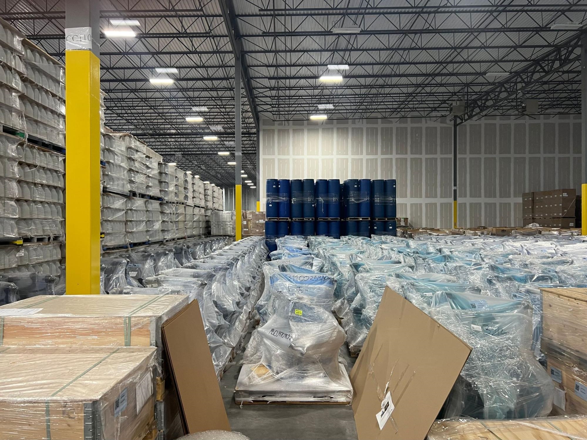 A wide warehouse aisle lined with tall shelving and stacked, shrink-wrapped pallets, with cardboard boxes in the foreground and drums stored on a middle rack.
