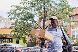 A delivery worker is carrying a cardboard box package while walking outside on a city street.