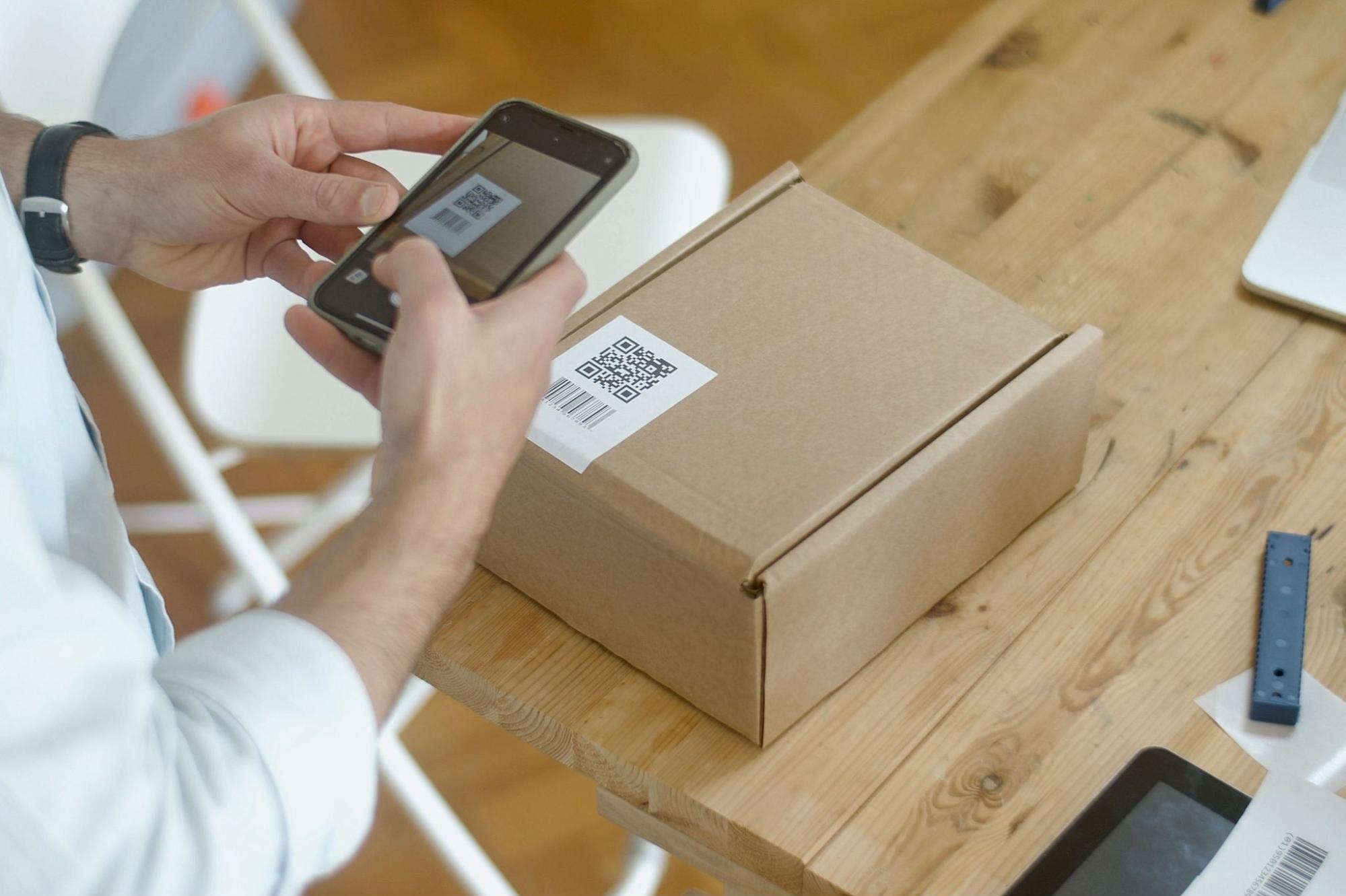 A person scans a QR code on a cardboard shipping box with a smartphone at a wooden table.