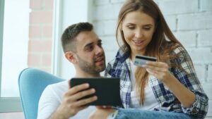 Two people look at a digital tablet together indoors. One is sitting on the other’s lap while holding a blue credit card.