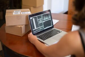 A person works on a laptop at a wooden table next to a stack of shipping boxes.