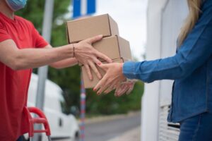 A delivery worker in a red shirt and face mask hands a stack of cardboard boxes to a person in a denim jacket, with a white delivery van parked nearby.